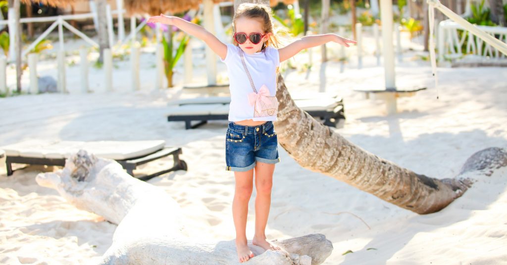 Niña descalza posando con los brazos abiertos y gafas de sol en una playa.